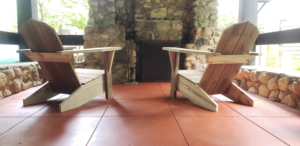 The backs of two unpainted Adirondack chairs facing a stone fireplace in an outdoor shelter. The concrete floor is painted grand Canyon red.